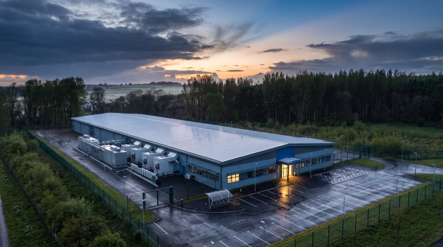 Aerial view at dusk of a single-story data center with rooftop cooling plant, an empty wet parking lot, and a security fence, set among trees and farmland under a clearing sky.